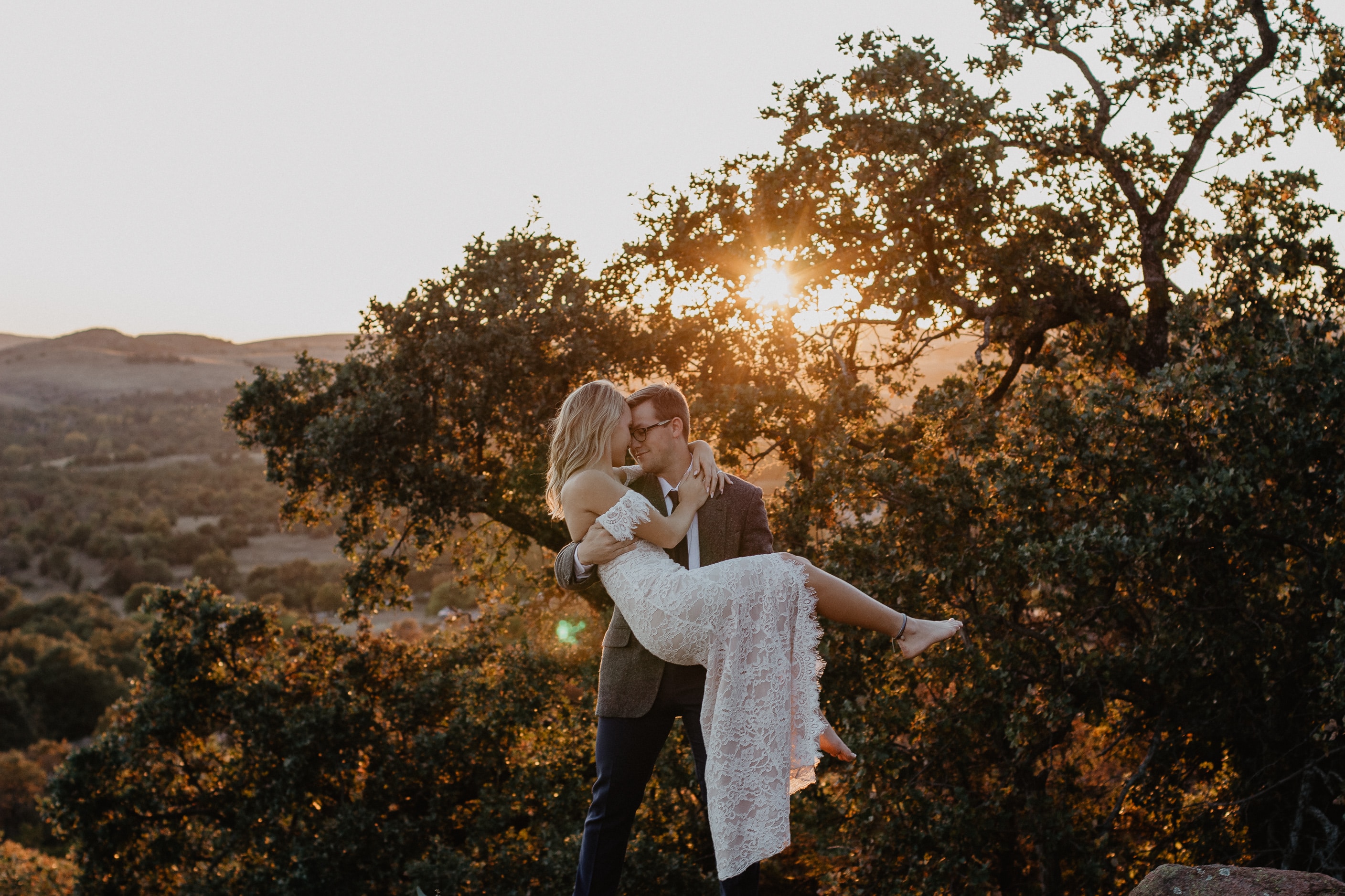 groom picking up his bride at sunset in front of a tree in a field