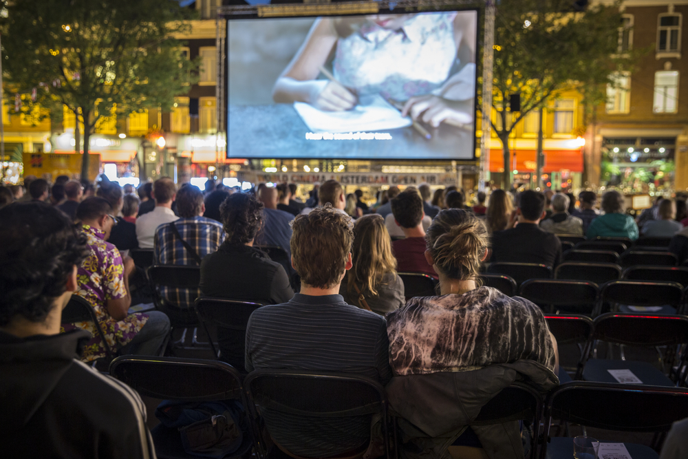 Families watch a movie on a large screen in a park.