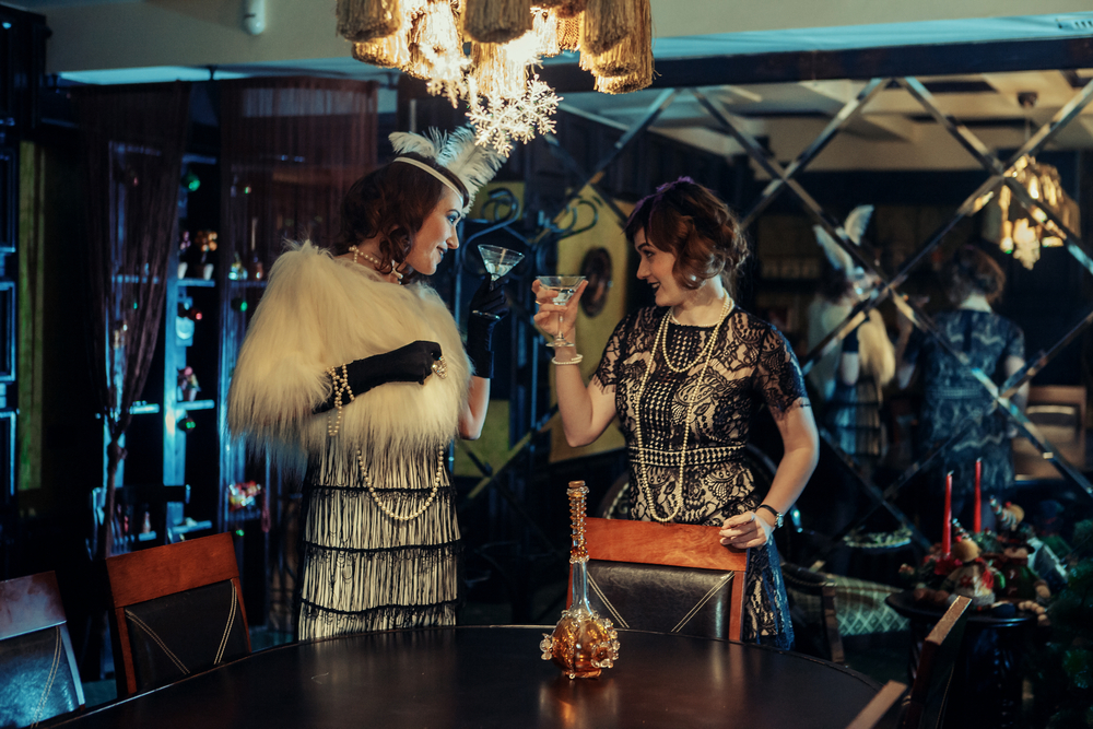 Two women dressed in 1920's American attire cheer in a dark lit bar.